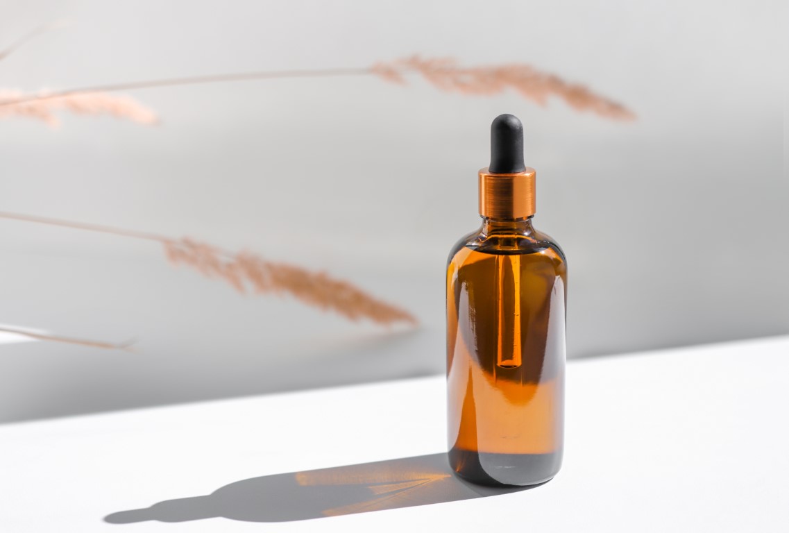 Amber glass bottle with cosmetic body oil on a white background with sunbeams and dry plants. Dropper serum, natural product, eco concept.